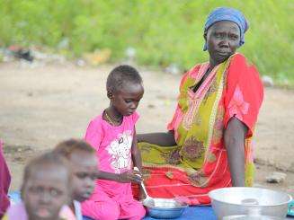 A mother sits outside with her children—MSF patients—as they prepare meals at thet Renk Civil Hospital