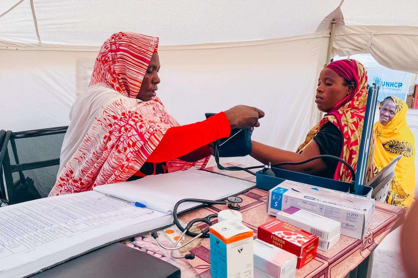 A female MSF medic takes the blood pressure of a female patient inside a white tent, other women wait outside and there are supplies and medication on the table in front of them. 