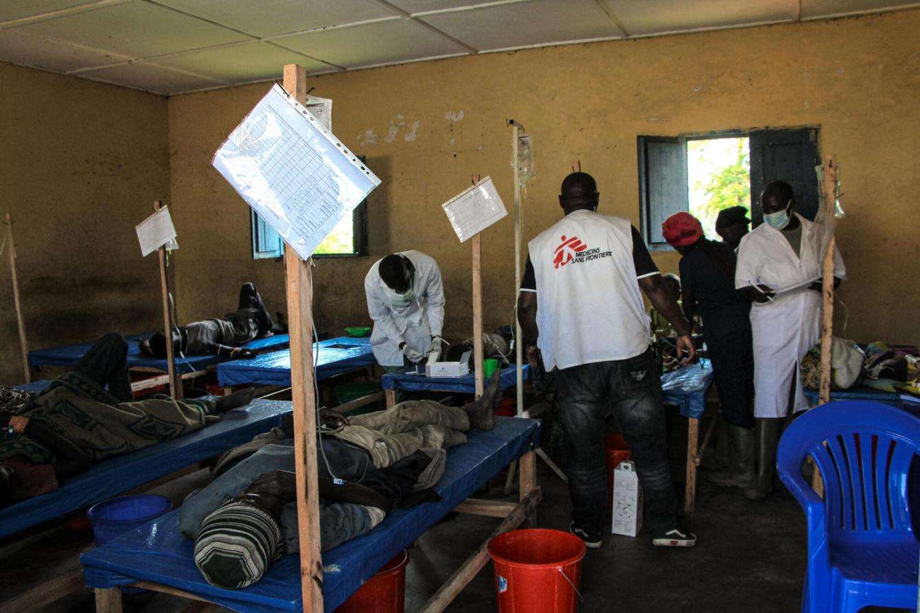 Inside of a former school with yellow walls and a small window, beds are set up and patients are laying on them and MSF doctors are gathered around one of the bed providing care.