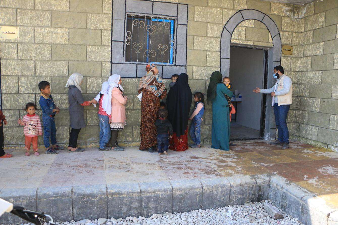 A long line of patients are standing outside of a stone building being directed inside by a medic. 