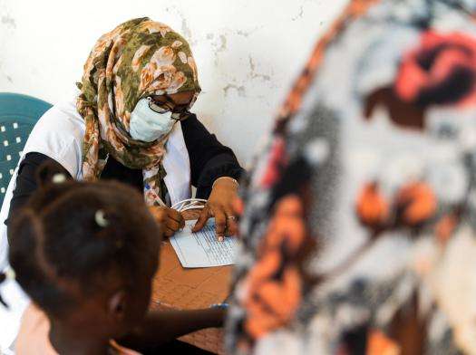 An MSF doctor in surgical mask, headscarf, and white vest writes a note at a mobile clinic in Wad Madani, Sudan.