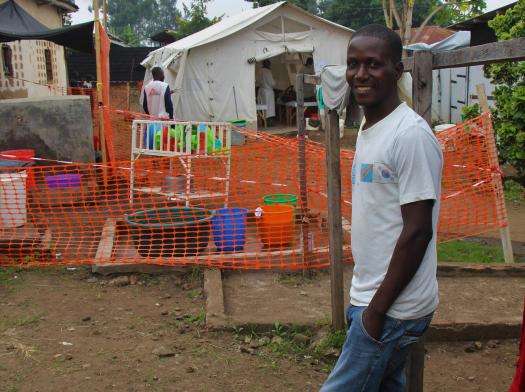 A man in a white T-shirt and jeans is walking outside of the one of MSF's cholera centers, smiling at the camera.