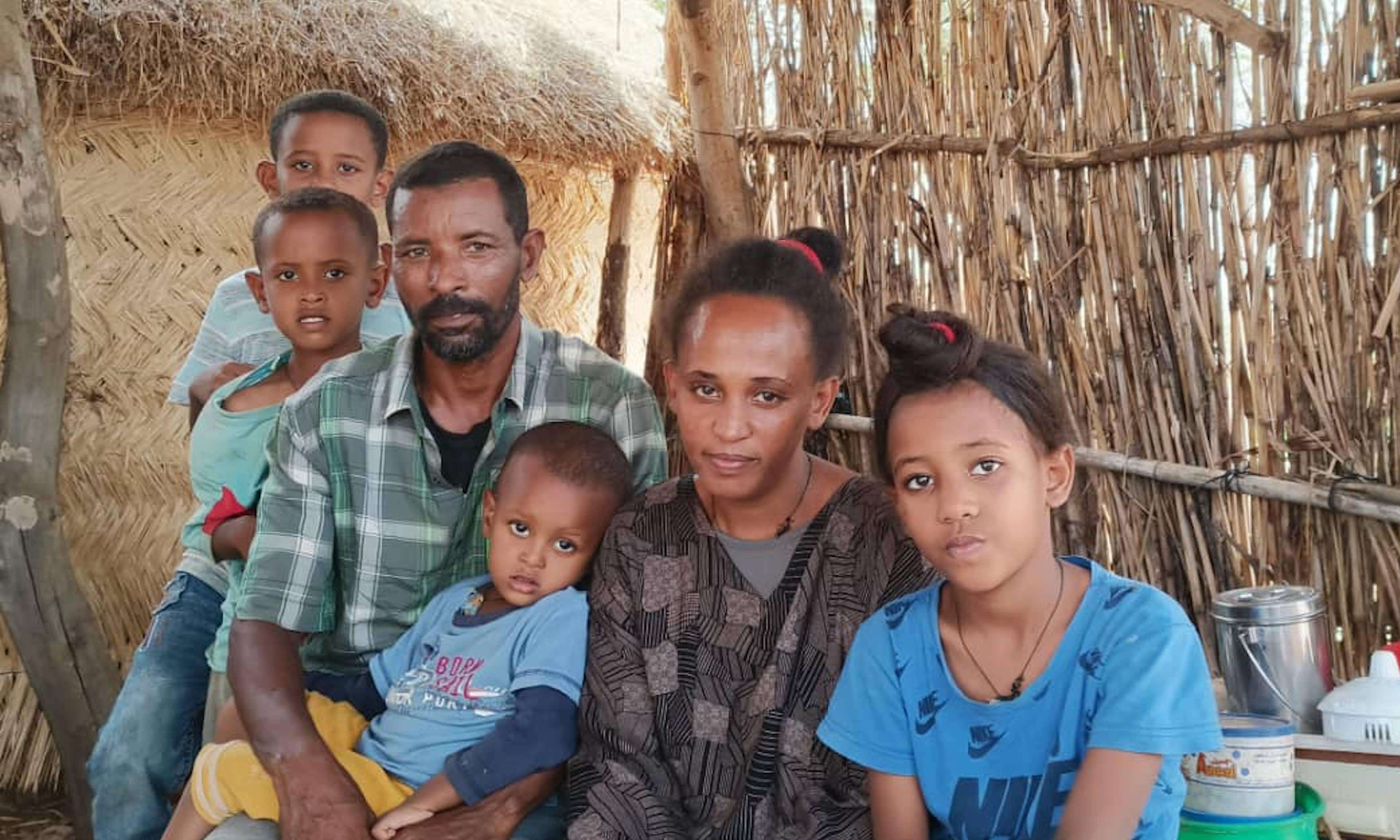 A family of four kids and a mother and father sitting under a thatched roof looking at the camera. 