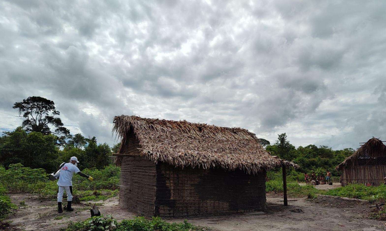 An MSF staff member is walking through a rural area to a hut with a thatched roof. 