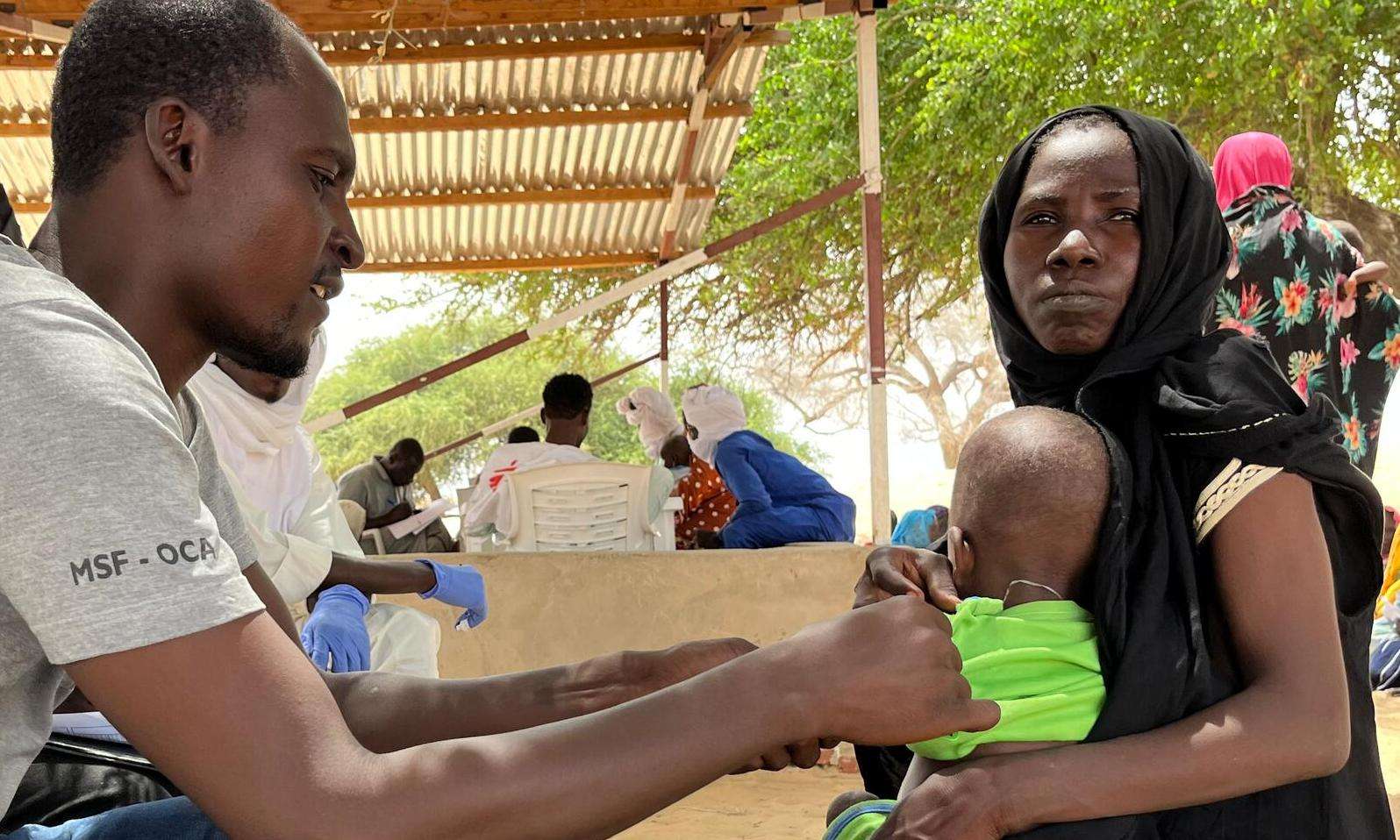 A man in a T-shirt with the MSF emblem is treating a child sitting in the mother's lap.