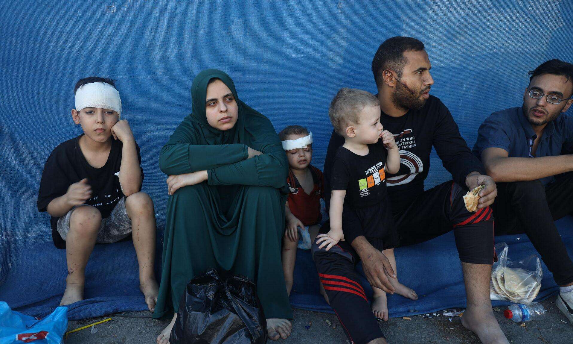 Wounded and displaced Palestinians in Gaza sit in front of a blue wall 