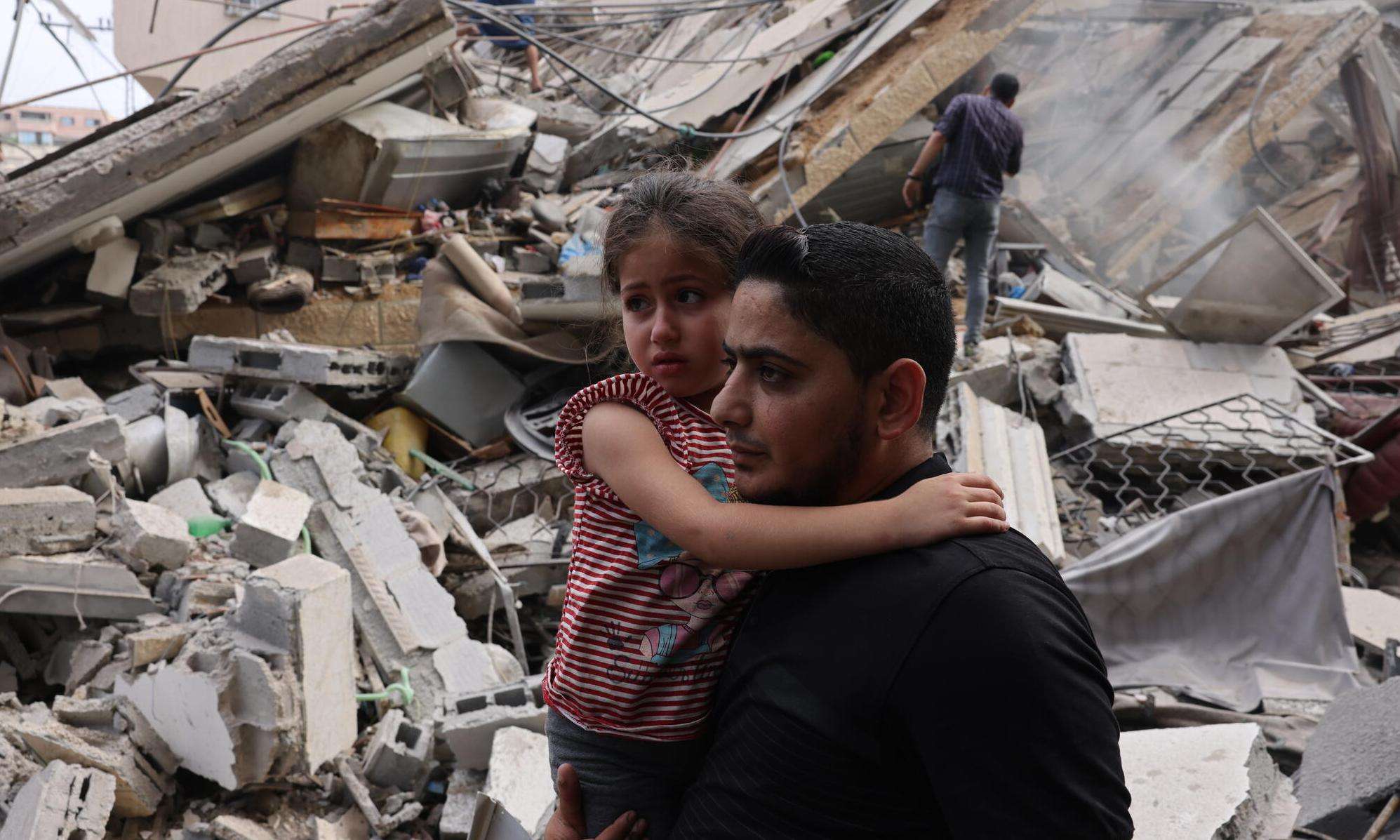 A man holding a child with wreckage from Israeli bombardment in Gaza behind them.