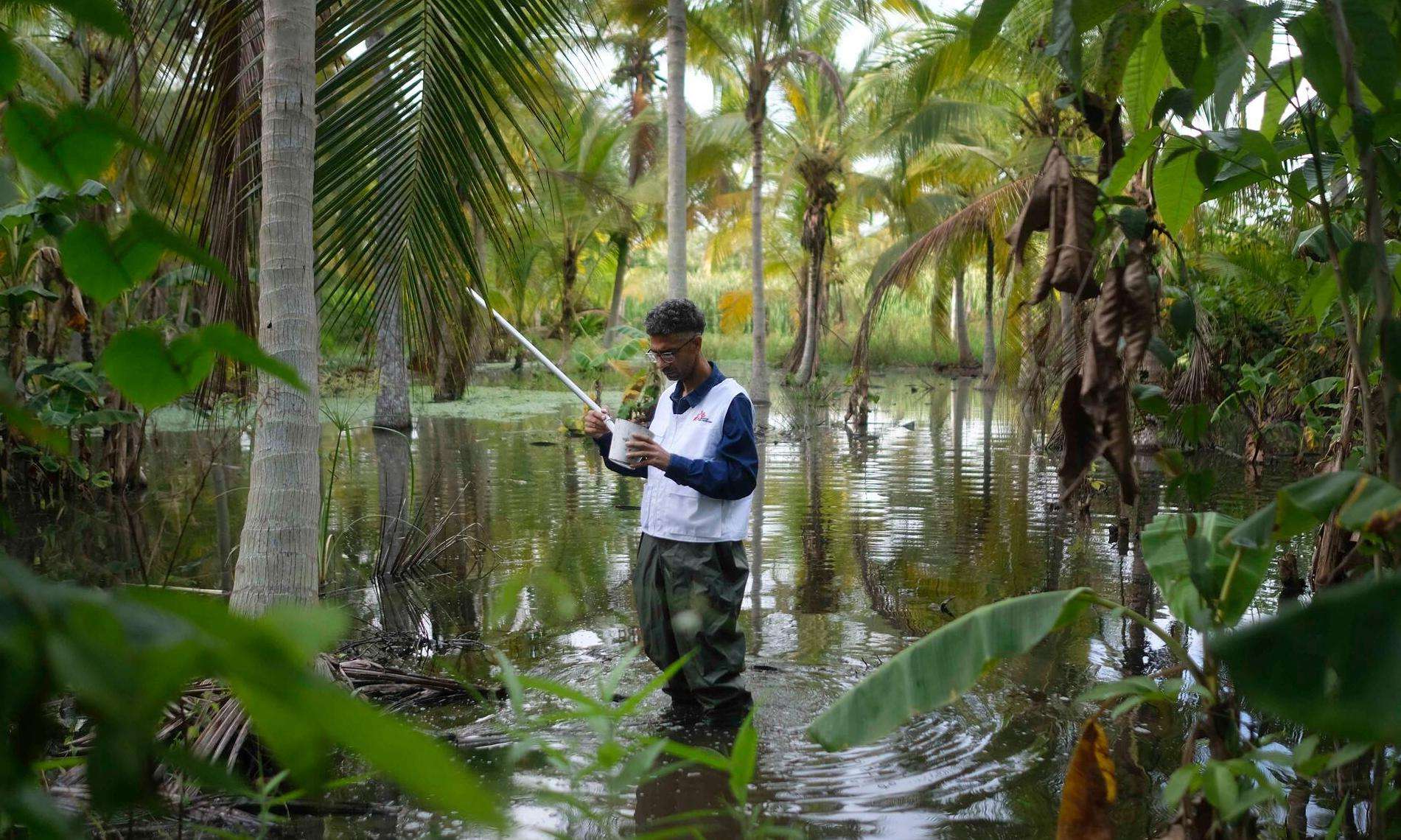 An MSF staff member wearing a white vest stands in the middle of floodwater surrounded by palm trees in Venezuela.
