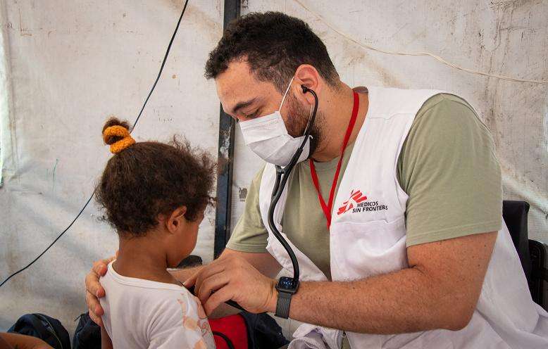 A doctor wearing a mask and MSF vest holds a stethoscope to a young girl's chest