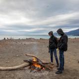 Two people standing next to a small campfire on a beach.