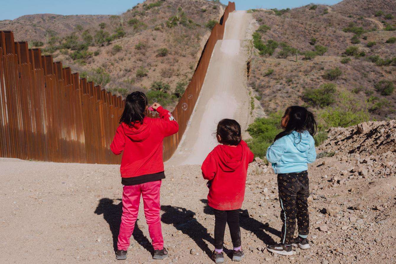 Children standing along the border wall between the United States and Mexico.