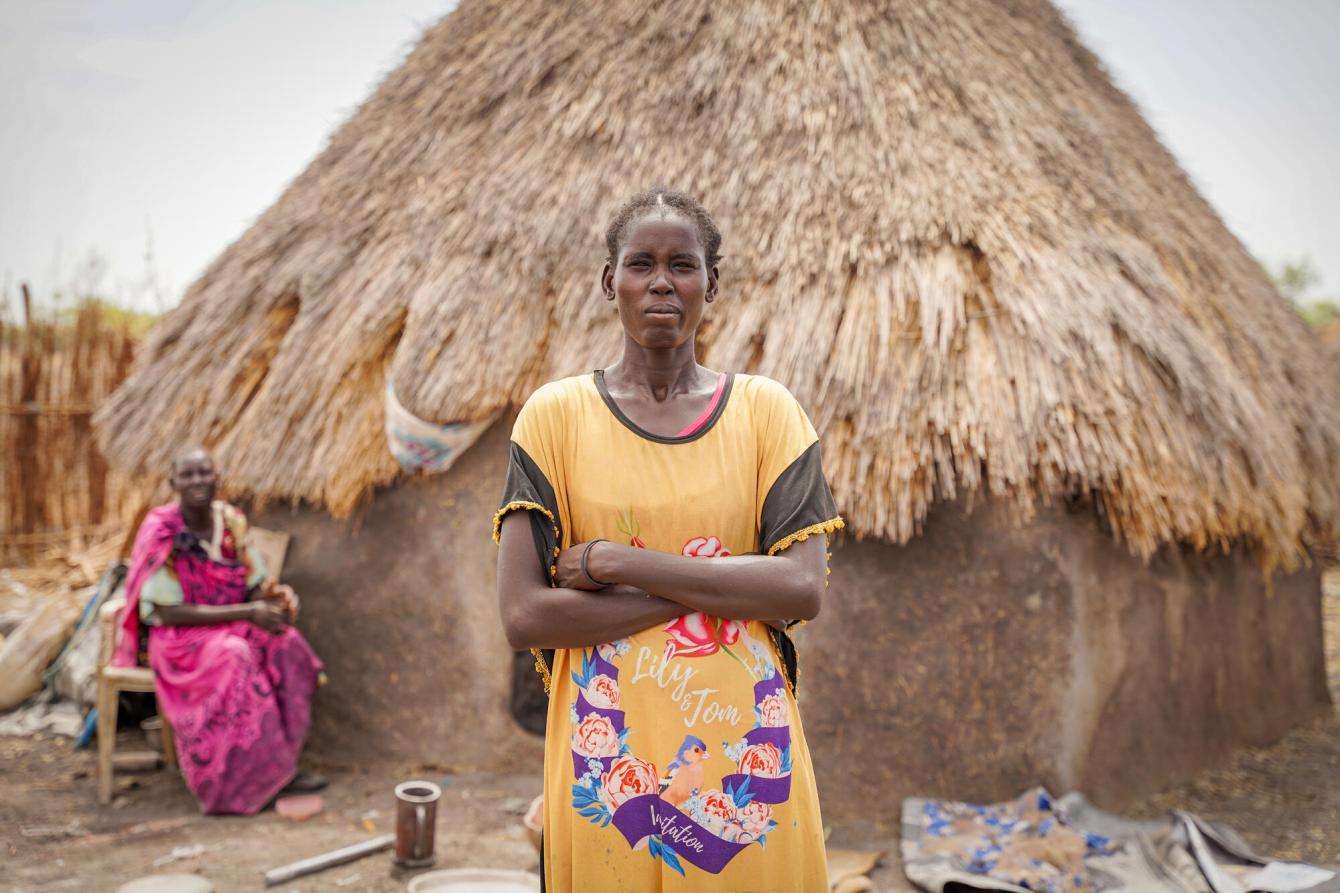 A woman in orange dress stands with arms crossed in front of hut in Aree, South Sudan under cloudy sky. 