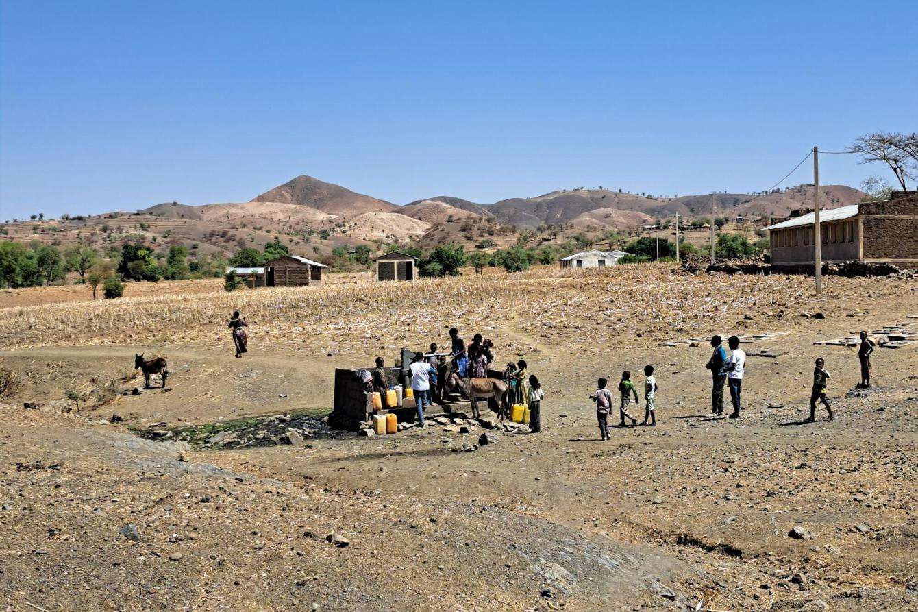 A group of people gather at a water hand pump in a field in Tigray, Ethiopia, with low mountains in the distance.