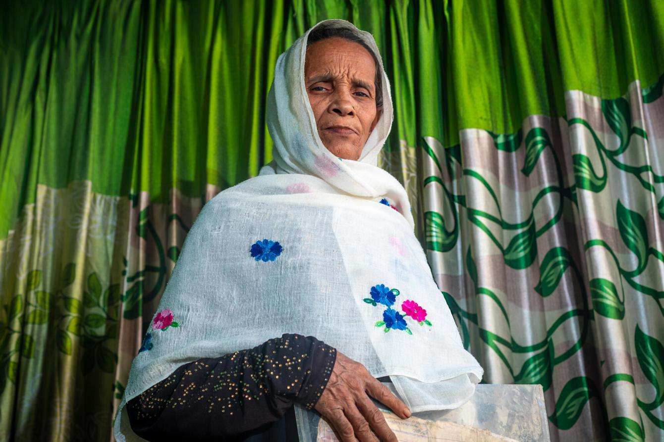 An older Rohingya woman, Melua, wears a white scarf and stands against a green curtain in Cox's Bazar, Bangladesh. 