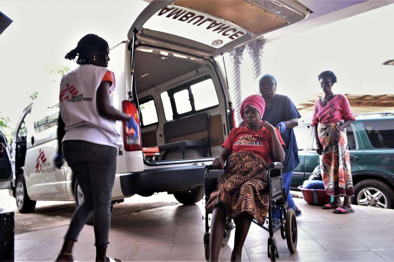 An MSF patient in a wheelchair outside of an MSF ambulance in SIerra Leone, where she is greeted by MSF staff.
