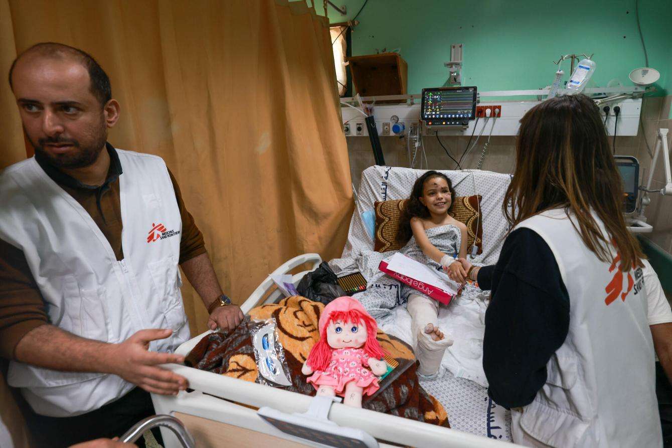 A young girl lies in a hospital bed greeting MSF staff at Al Aqsa Hospital in Gaza.