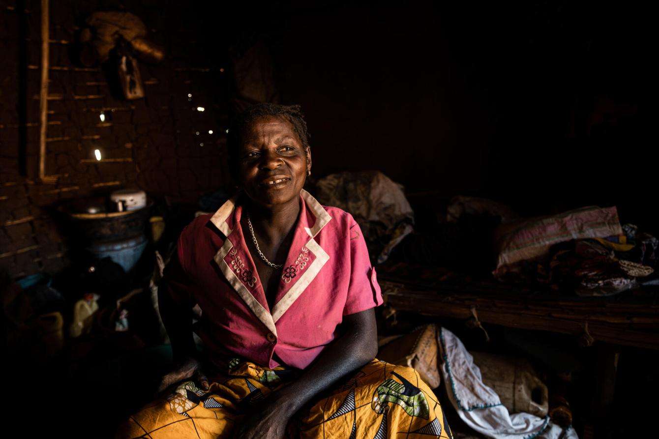Portrait of a woman in a pink shirt in a hut in Dhenja, Democratic Republic of Congo, where she hosts other displaced people.