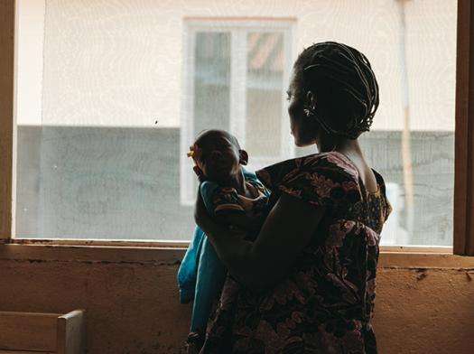 A woman holding a child with back turned looking out of an open window in Central African Republic.