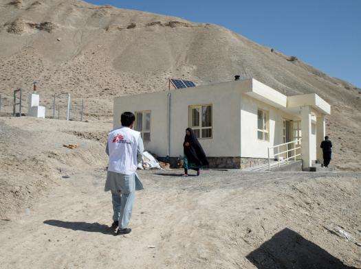 An MSF staff member in a white vest walks toward an MSF clinic and a woman in black veil in Afghanistan.