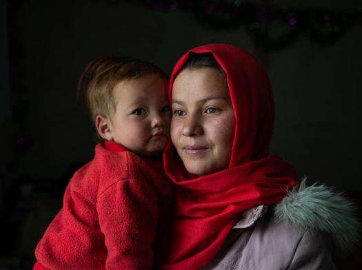 An Afghan young woman in a red headscarf holds her child in a red shirt against a dark background.