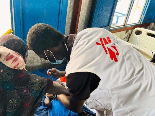 MSF aid worker in white vest bends over a hospital bed treating a patient with a blood compress. 