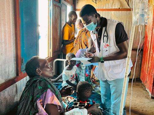 A man in an MSF vest stands over a patient taking notes on his clipboard. 