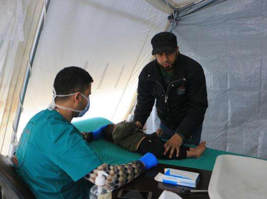 A man stands over a child that is laying on a hospital bed while a medic examines the child. This is all taking place inside of a tented medical space.