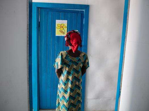 A woman in a green dress and red headscarf stands in front of a blue door at MSF's Tumaini clinic in Democratic Republic of Congo.