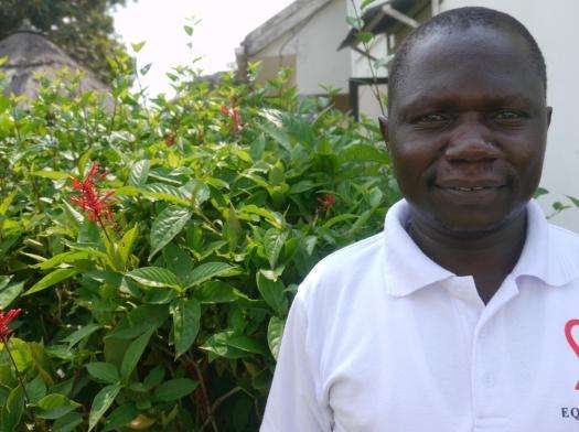 A man who is a former MSF patients in the Arua, Uganda, stands in front of a flowering bush, wearing a white T-shirt and smiling.