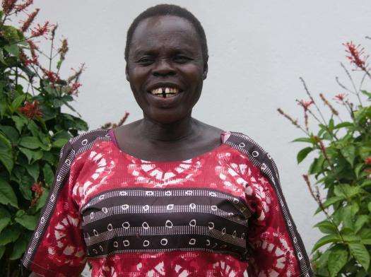 A woman who was an MSF patient in Arua smiles and stands between flower bushes.