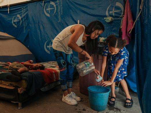 A displaced mother and child fill a bucket of water in their tent in Venezuela.