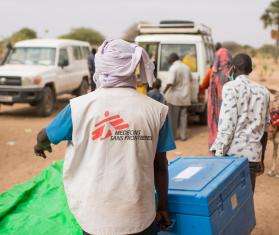 MSF aid worker in white vest with logo carries donations of supplies to vehicles in Chad