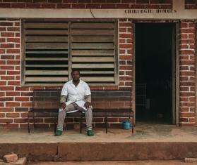 Dr. Sabio sits in white lab coat in front of the brick wall of MSF-supported Bakouma secondary hospital in Central African Republic.